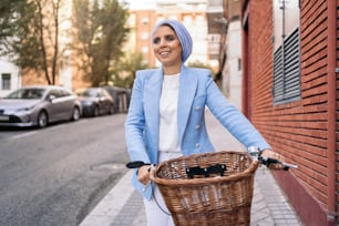 Imagen horizontal de una mujer musulmana vestida con un traje de luz azul y pantalones blancos caminando con su bicicleta