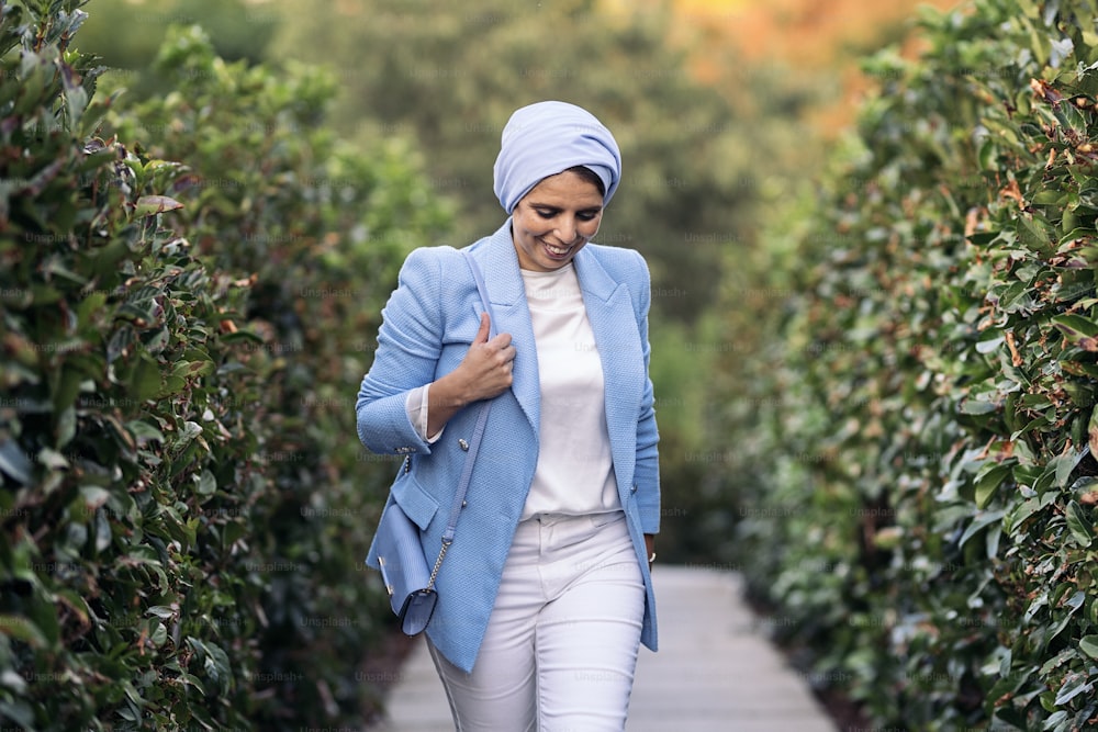 Front view of a smiling muslim woman walking among large bushes wearing a blue light suit and headscarf holding her purse on a pathway looking down in a sunny day.