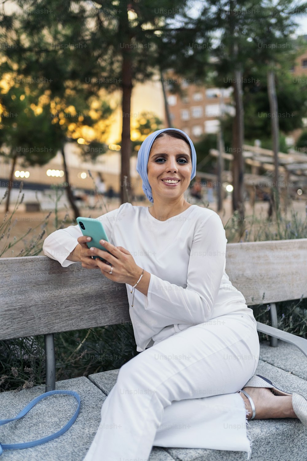 Front view of a muslim woman wearing a light blue headscarf sitting on a bench in the park using her phone.