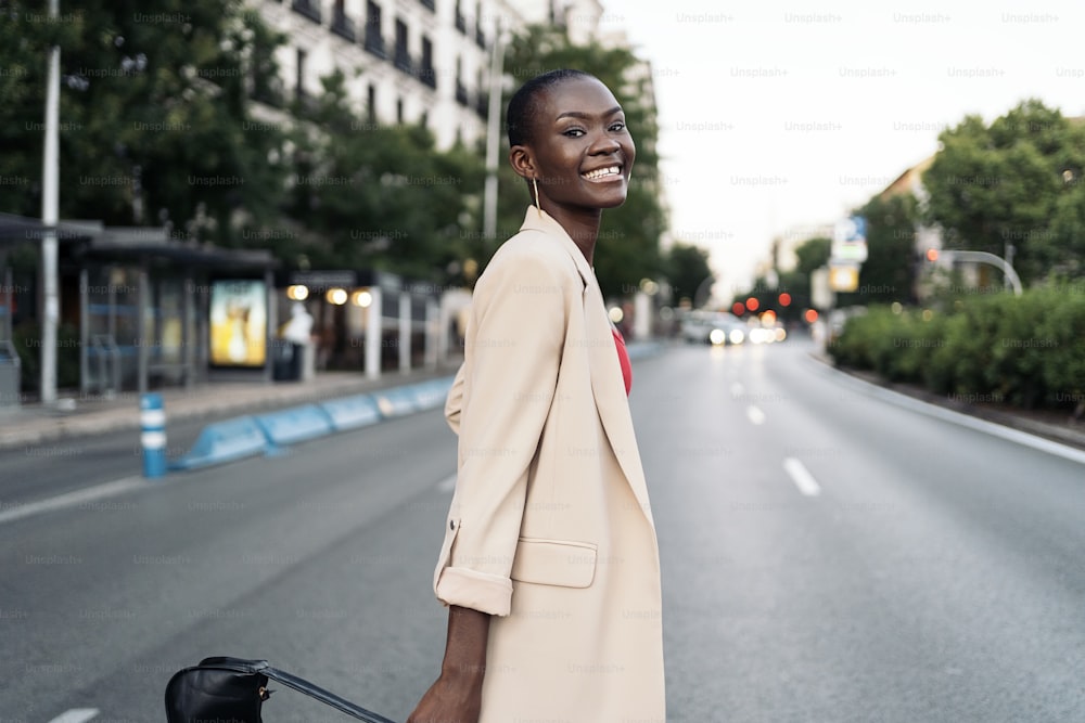 Mulher jovem adulta elegante com cabelo curto olhando para a câmera e sorrindo enquanto atravessa a rua na cidade