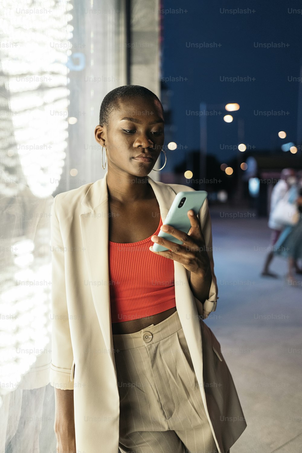 Porträt einer modischen jungen Frau mit kurzen Haaren, die ihren Körper nachts an ein Schaufenster lehnt, während sie ihr Telefon benutzt