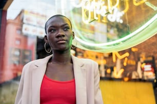 Ritratto di una donna adulta afroamericana alla moda in piedi e distogliendo lo sguardo con l'insegna al neon dietro in città
