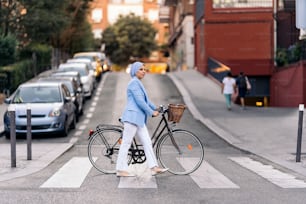 Vertical image of a muslim woman wearing a blue light suit and white pants walking with her bicycle crossing the street looking to a side