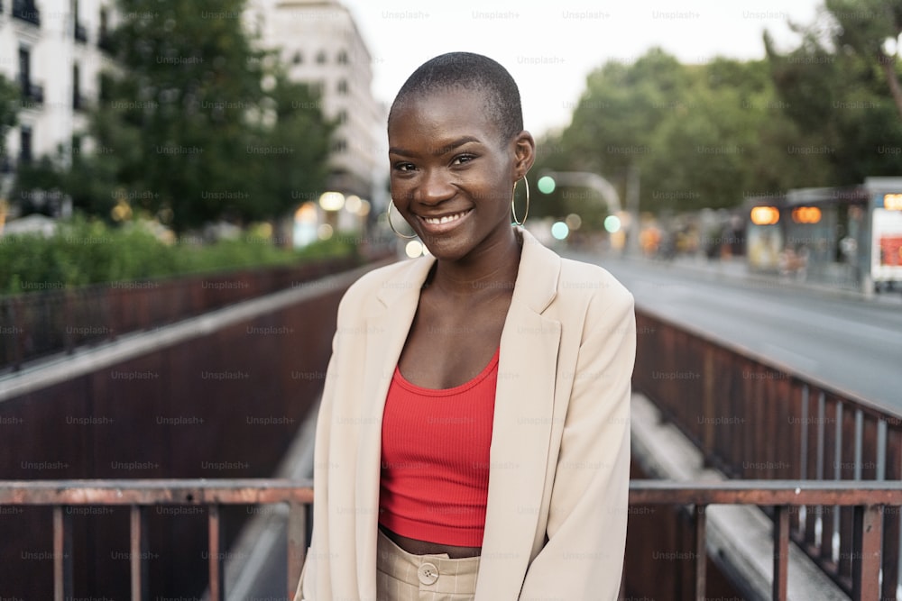 Mulher jovem afro-americana sorridente com cabelo curto em pé e olhando para a câmera em uma passarela na cidade