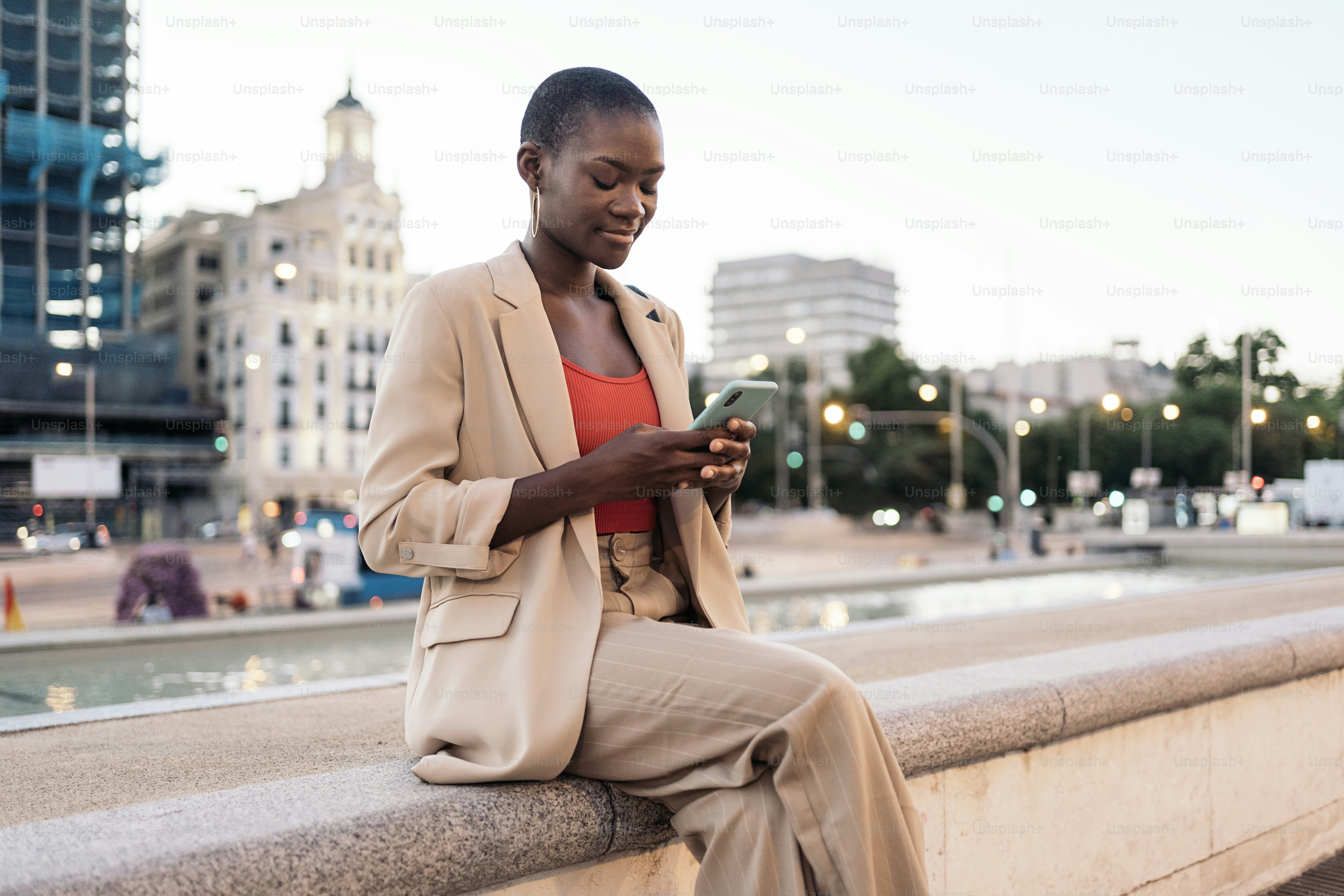 Vista a figura intera di una donna alla moda seduta sul bordo di una fontana che usa il suo telefono