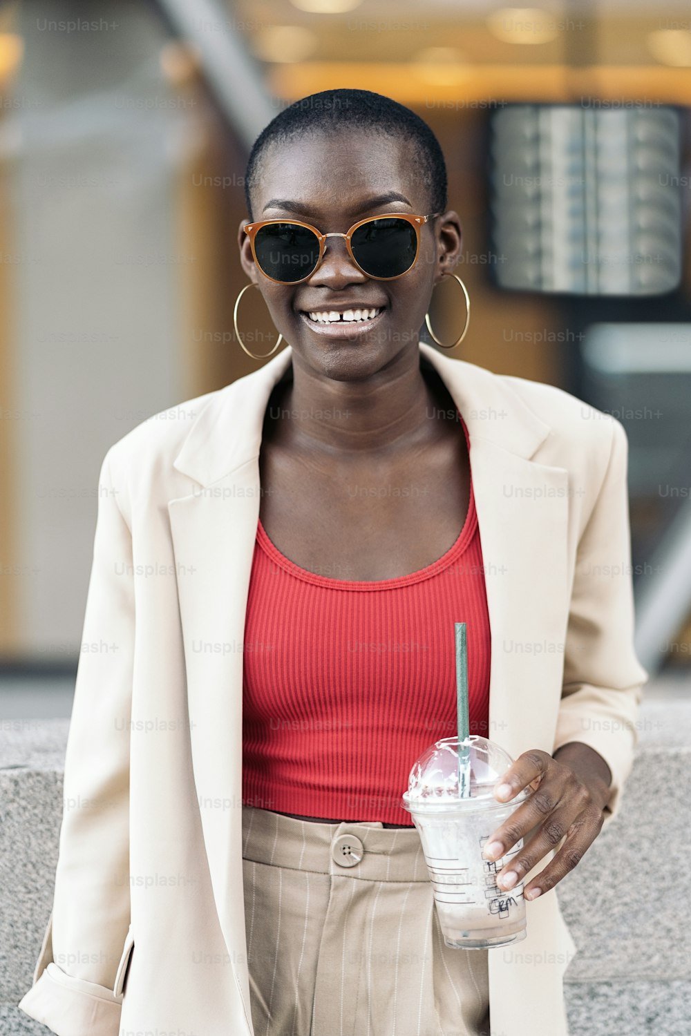 Vorderansicht einer eleganten, fröhlichen afroamerikanischen erwachsenen Frau mit kurzen Haaren und Sonnenbrille, die einen Milchshake hält und in die Kamera schaut