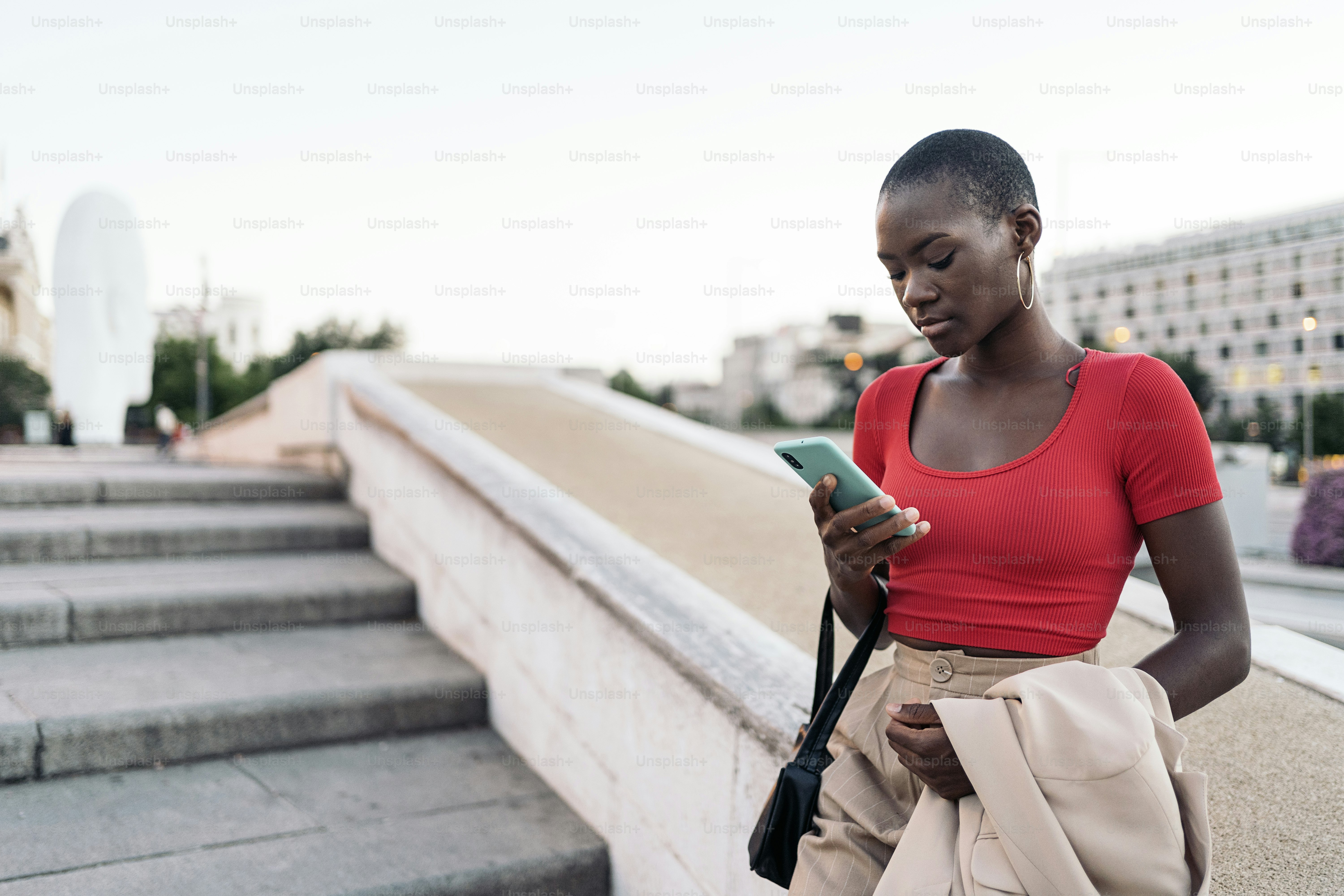 Elegant young adult woman with short hair using phone while holding her jacket and bag on outdoor stairs in the city.