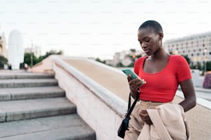 Elegant young adult woman with short hair using phone while holding her jacket and bag on outdoor stairs in the city.
