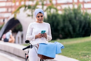 하늘색 머리 스카프를 두른 무슬림 여성이 공원에서 자전거를 들고 서 있는 동안 휴대전화를 사용하는 모습