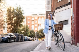 Horizontal image of a muslim woman wearing a blue light suit and white pants walking with her bicycle