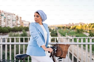 Side view of an unsmiling muslim woman standing with her bike on a footbridge looking to a side in a sunny day