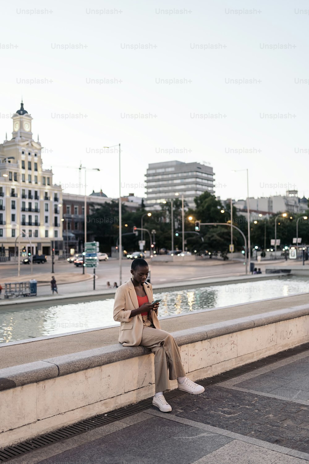 Full length view of a fashionable woman sitting on the edge of a fountain using her phone