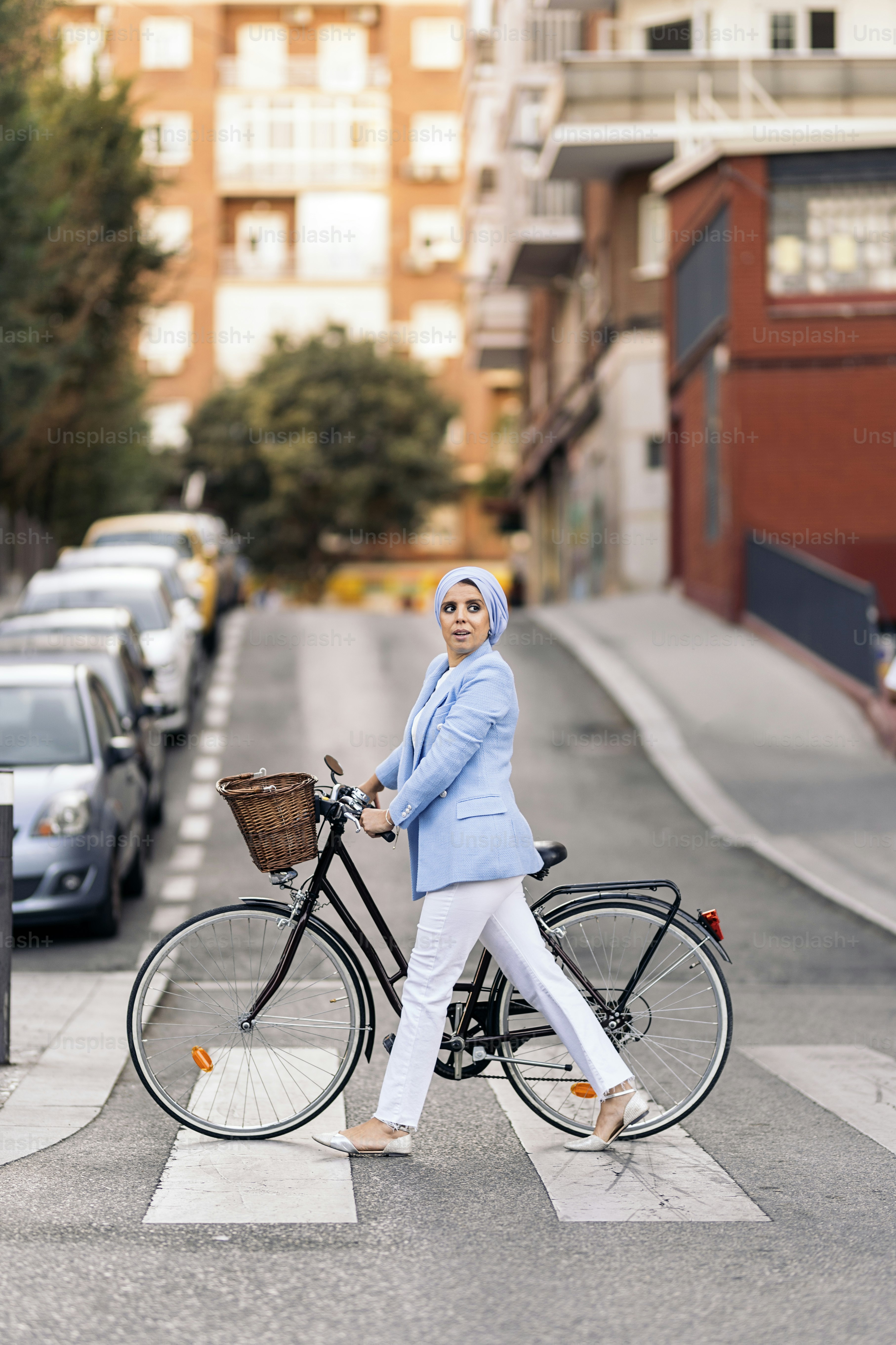 Vertikales Bild einer muslimischen Frau in blauem Anzug und weißer Hose, die mit ihrem Fahrrad die Straße überquert und zur Seite schaut