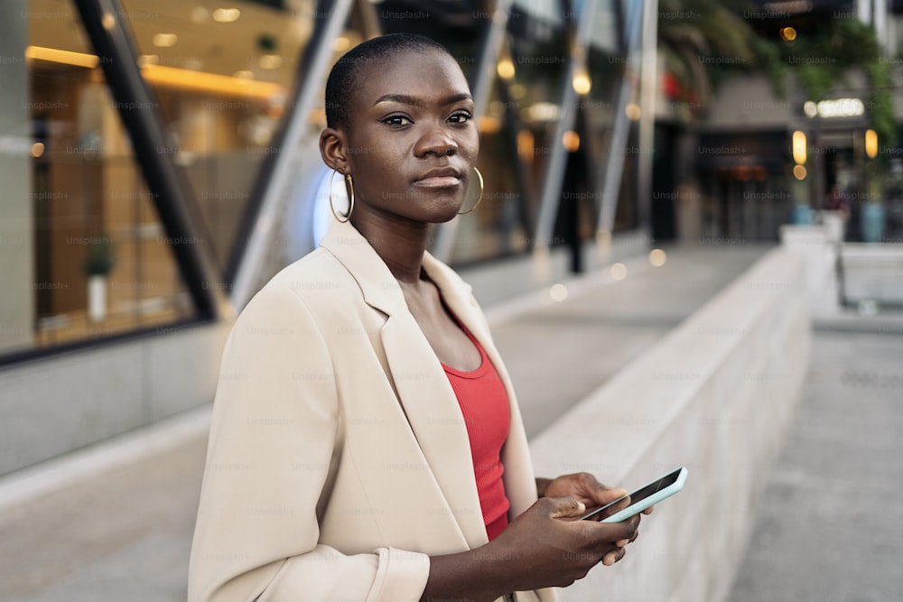 Vista laterale di una giovane donna afroamericana adulta con i capelli corti in città usando il suo telefono e guardando la telecamera