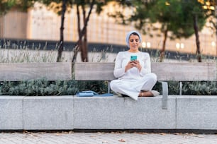Front view of a muslim woman wearing a light blue headscarf sitting on a bench in the park using her phone.