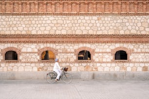 Vista lateral de una mujer musulmana montando su bicicleta en la acera con una gran pared de ladrillo con ventanas en un día soleado.