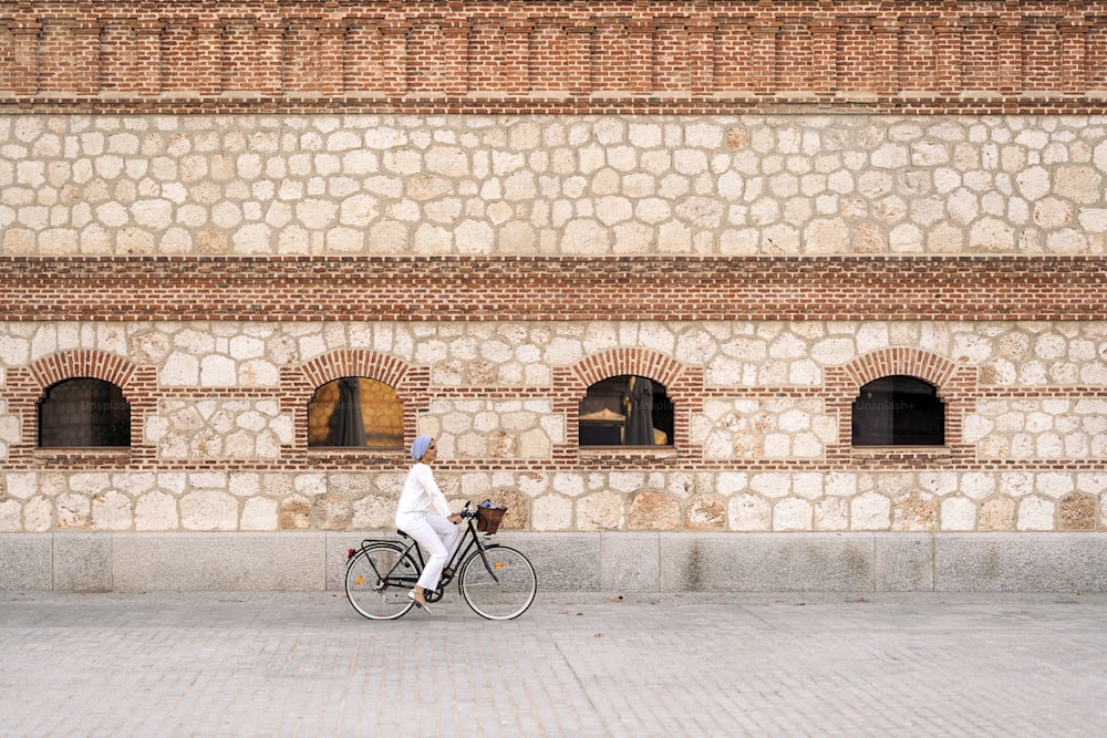 Seitenansicht einer muslimischen Frau, die an einem sonnigen Tag mit ihrem Fahrrad auf dem Bürgersteig mit einer großen Ziegelmauer mit Fenstern fährt.