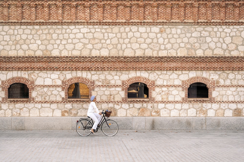 Side view of a muslim woman riding her bicycle on the sidewalk with a large brick wall with windows in a sunny day.