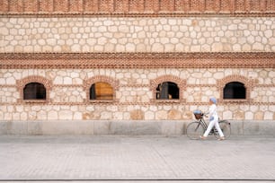 Vista lateral de uma mulher muçulmana segurando sua bicicleta na calçada com uma grande parede de tijolos com janelas em um dia ensolarado.