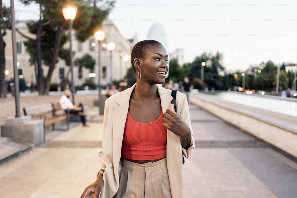 Front view of a fashionable african american young adult woman laughing while walking in the city at sunset