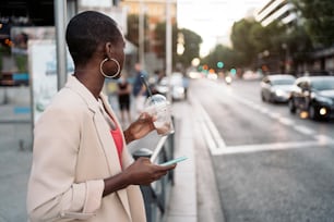 짧은 머리와 양복을 입은 젊은 아프리카계 미국인 여성이 전화기와 밀크셰이크를 들고 길을 건너기 위해 옆을 바라보며 기다리고 있다