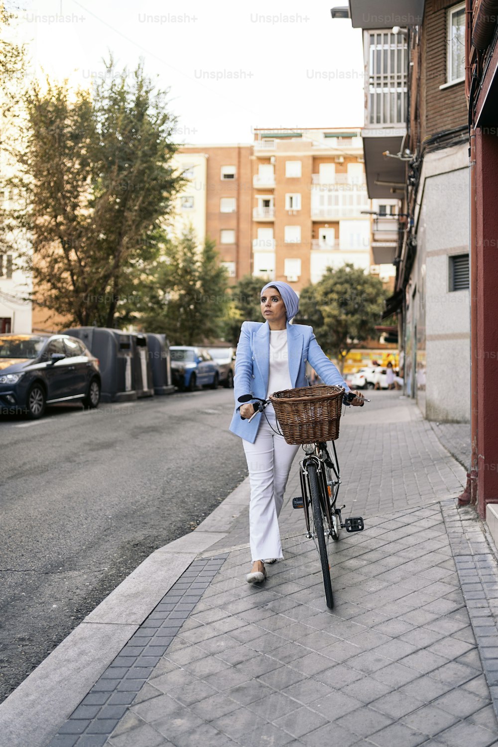 Imagen vertical de una mujer musulmana vestida con un traje de luz azul y pantalones blancos caminando con su bicicleta