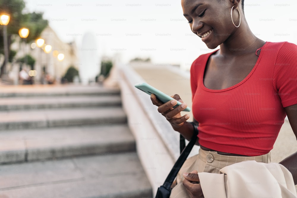 Nahaufnahme einer afroamerikanischen jungen erwachsenen Frau, die ihr Telefon benutzt, während sie ihre Jacke und Tasche auf einer Außentreppe in der Stadt hält