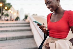 Primer plano de una mujer adulta joven afroamericana usando su teléfono mientras sostiene su chaqueta y bolso en las escaleras al aire libre de la ciudad