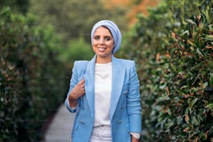 Vorderansicht einer lächelnden muslimischen Frau, die zwischen großen Büschen steht und einen Blaulichtanzug und ein Kopftuch trägt, ihre Handtasche auf einem Weg hält und an einem sonnigen Tag in die Kamera schaut.