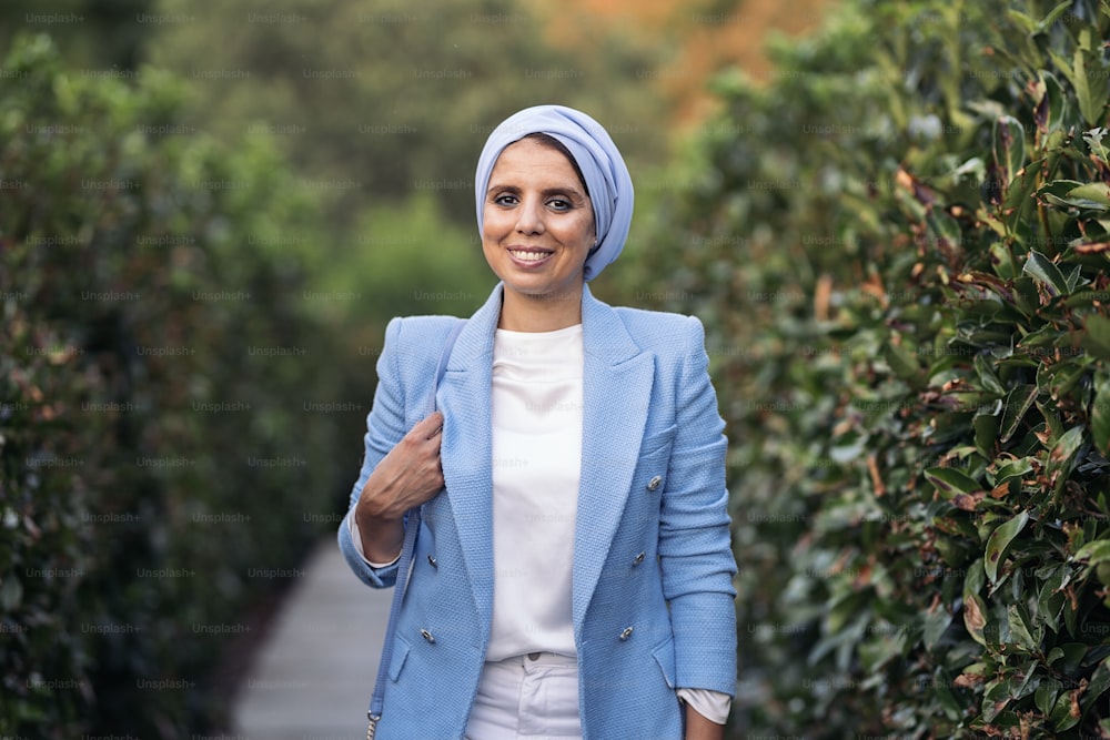 Front view of a smiling muslim woman standing among large bushes wearing a blue light suit and headscarf holding her purse on a pathway looking at camera in a sunny day.