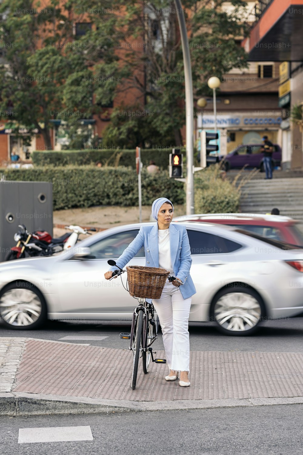 Vertical image of a muslim woman wearing a blue light suit and white pants walking with her bicycle crossing the street looking to a side