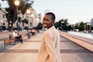 Rear view of a fashionable african american young adult woman walking forward and turning to look at the camera in the city at sunset