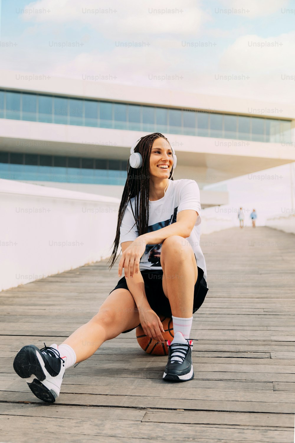 Mujer sonriente escuchando música con auriculares, ropa deportiva urbana sentada en una pelota de baloncesto. Con edificio de vidrio futurista en el fondo. Felicidad y deporte