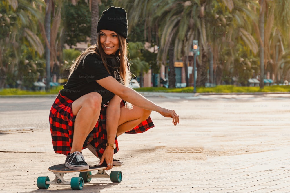Lächelnde junge Frau hockte sich mit dem Skateboard auf die Straße. Lateinamerikanische Frau übt auf einem Boulevard.