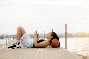 General shot of a basketball player resting, using her smart phone and headphones with her head on a ball in the harbor