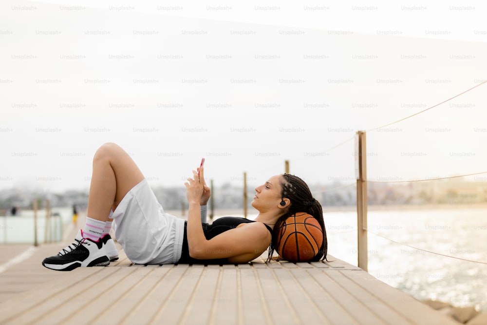 Scatto generale di una giocatrice di basket che riposa, usando il suo smartphone e le cuffie con la testa su una palla nel porto