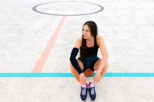 Müde junge Sportlerin sitzt mit Basketball auf dem Sportplatz
