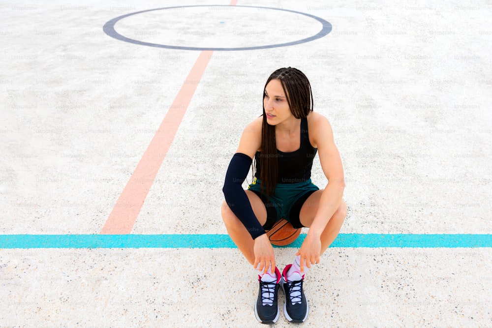 Giovane donna sportiva stanca che si siede con la pallacanestro al campo sportivo
