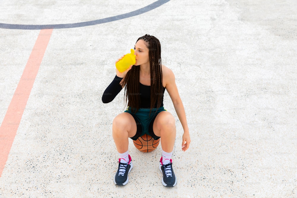 Scatto visto dall'alto di una giocatrice di basket seduta su una palla e bevendo acqua sul campo. Idratazione e riposo