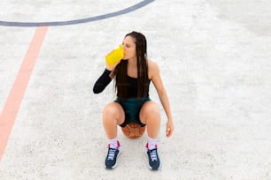 Aufnahme von oben einer Basketballspielerin, die auf einem Ball sitzt und Wasser auf dem Platz trinkt. Flüssigkeitszufuhr und Ruhe