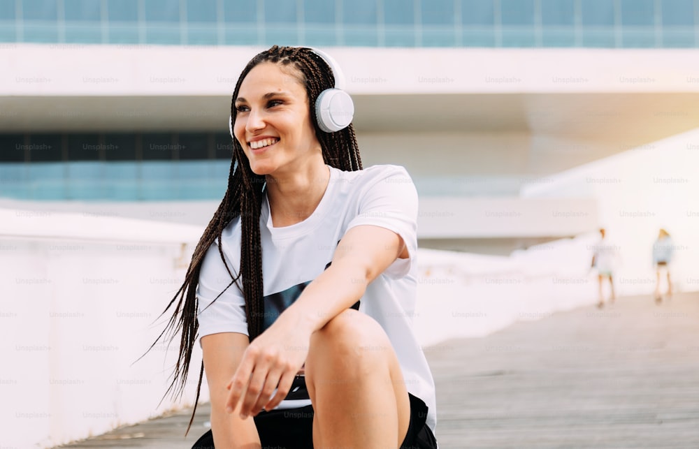 Mujer sonriente con trenzas en el pelo y auriculares escuchando música en su tiempo mirando al horizonte. Foto horizontal de estilo de vida. Chica de raza mixta