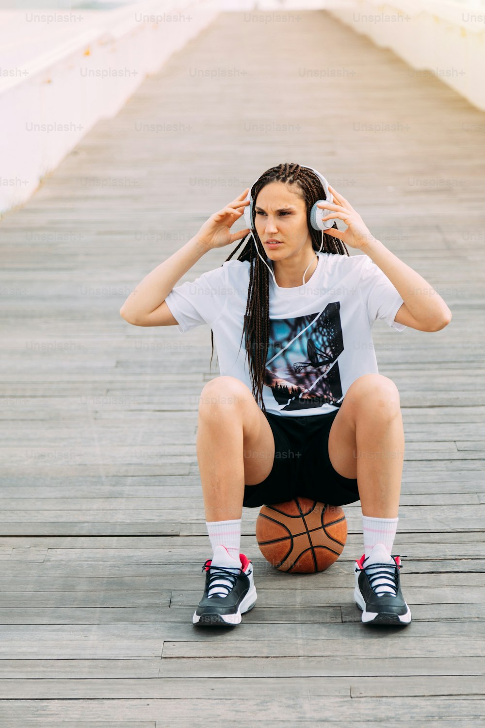 Fille métisse avec des tresses écoutant de la musique avec ses écouteurs, assise sur un ballon de basket. Audio, sport et personnes réelles