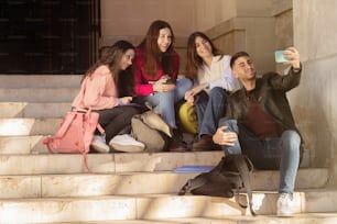 Group of friends taking a selfie sitting on the stairs. Four students at the university. Technology, academic classes. Leisure time