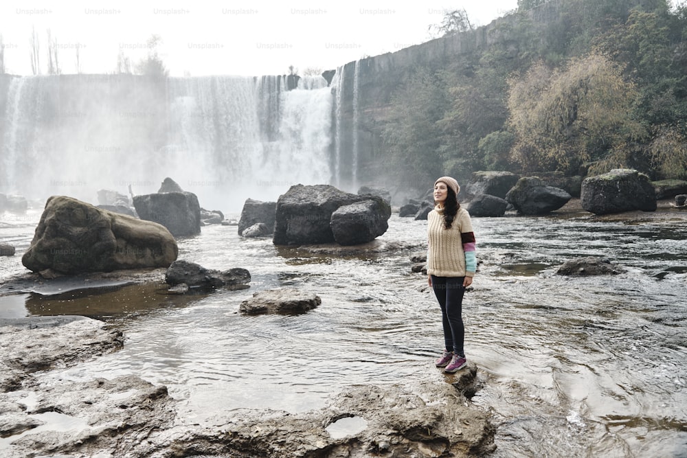 Jeune femme latine debout au bord d’une rivière avec une cascade en arrière-plan