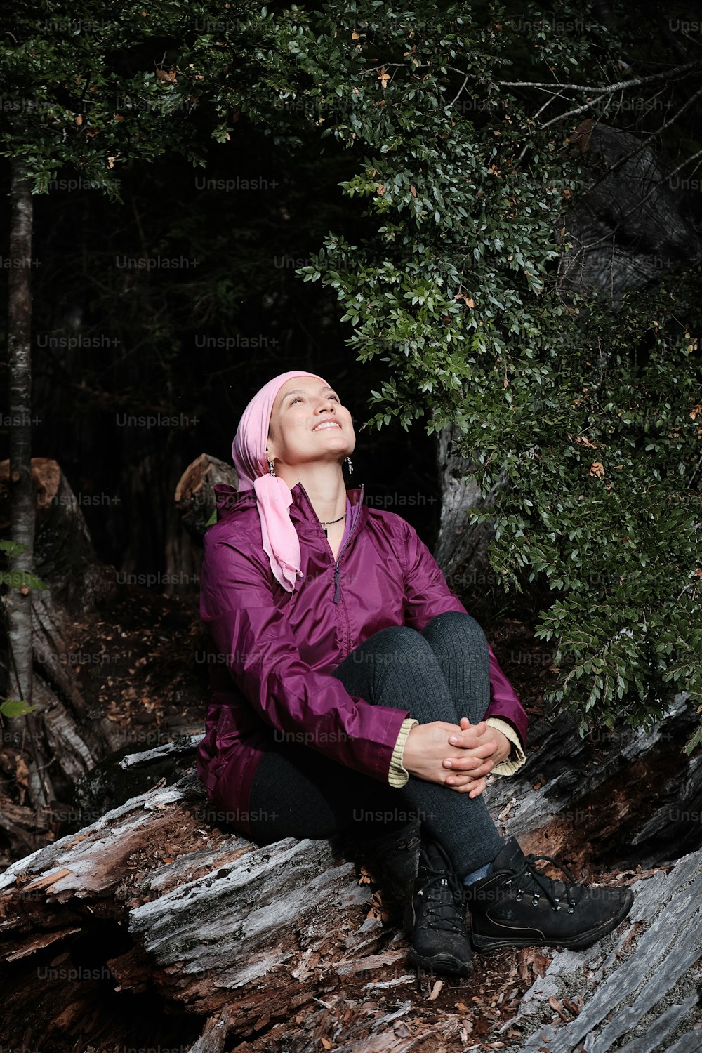 Vertical portrait of a young woman fighting cancer looking up to the sky and giving thanks for being alive sitting on a log of wood in the middle of the forest