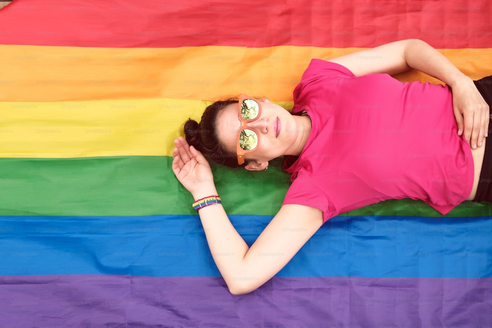 Frau mit Sonnenbrille, die auf einer Regenbogenflagge auf dem Boden liegt, die sexuelle Vielfalt darstellt. LGBTQ-Community-Konzept