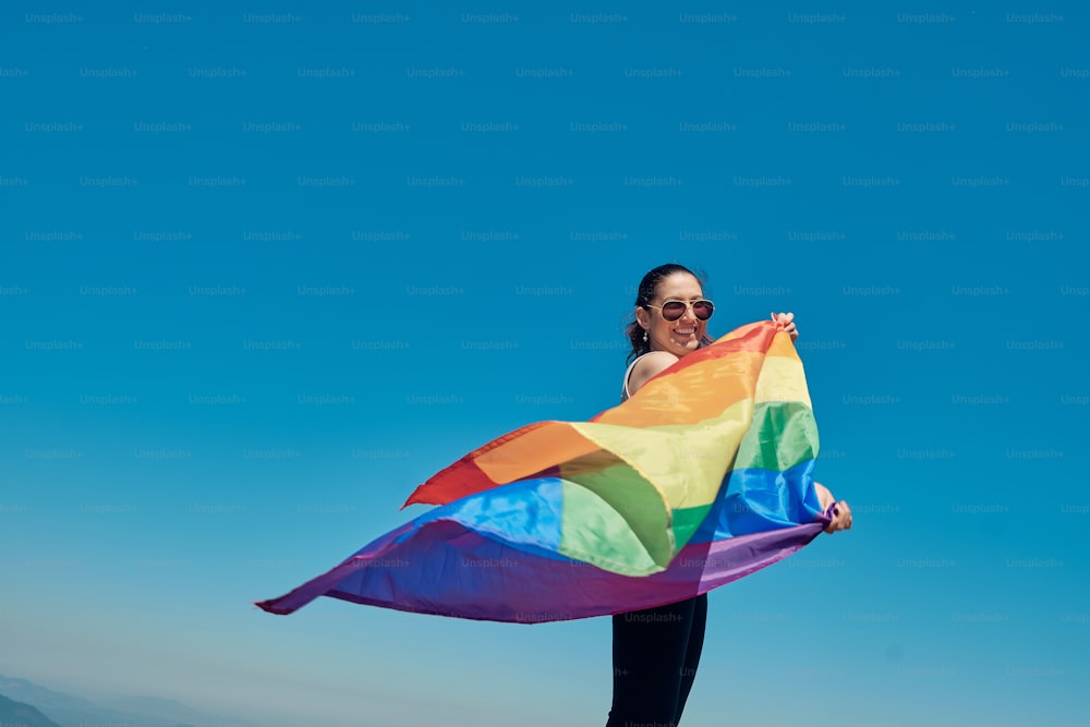 Young woman wearing sunglasses raises the pride flag over a blue sky background.