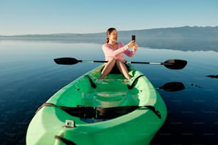 Mujer joven y feliz sentada en un kayak haciendo un video con su teléfono celular en medio de un tranquilo lago azul.