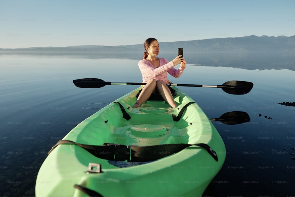 Donna giovane e felice seduta in un kayak facendo un video con il suo cellulare nel mezzo di un lago blu calmo.