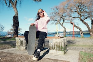 Cute girl sitting and leaning her hand on a skateboard. In the background the blue sky and the park. Concept of sports lifestyle and street culture